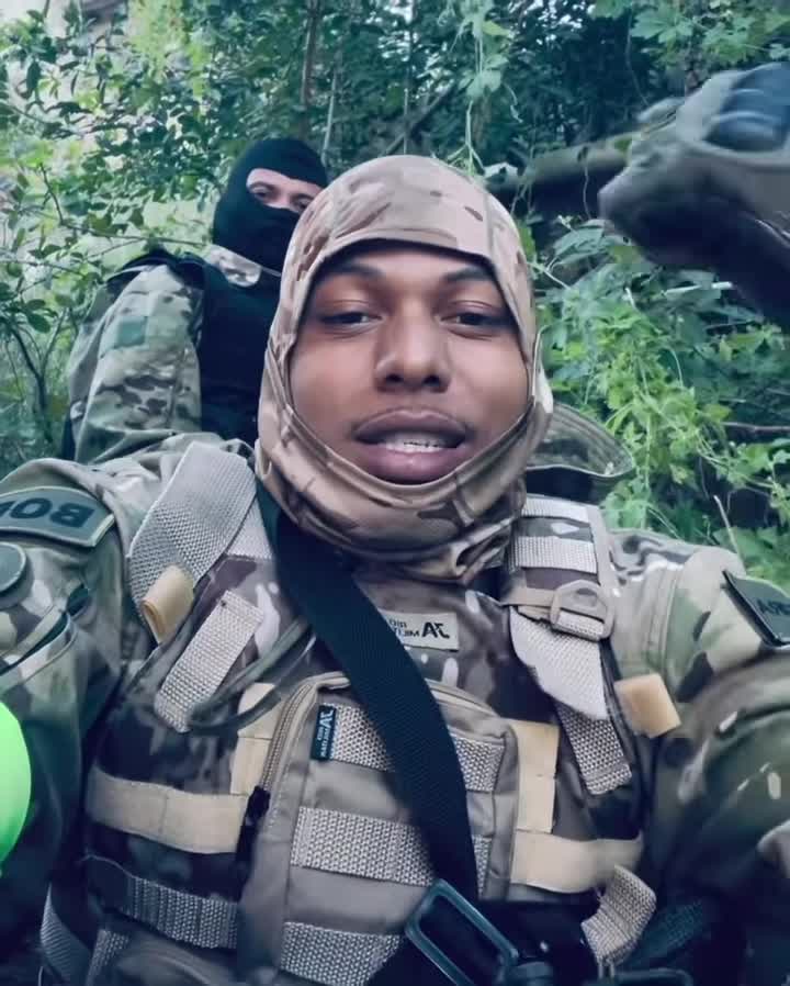 Homem fala à câmera com outra pessoa de rosto coberto ao fundo